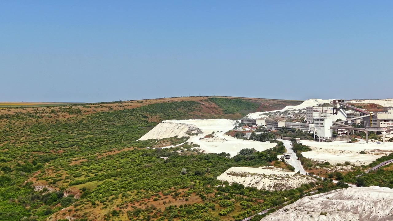 Grande área industrial com pilhas de mineração brancas e campos verdes ao fundo, em um dia ensolarado.