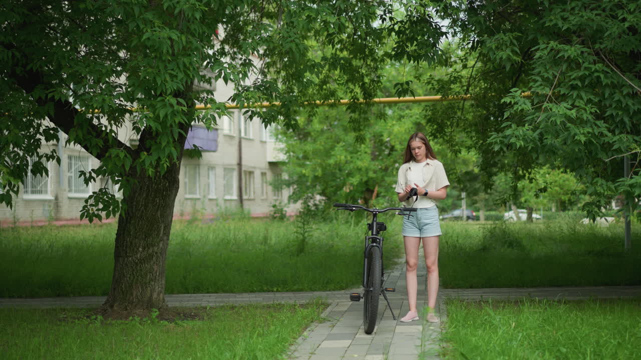 женщина держит черные перчатки, приближается к припаркованному велосипеду на асфальтированной дорожке, надевает перчатки в то время как смарт-часовки видны на правой руке, фон включает в себя пышную зелень, деревья и проезжающую машину на расстоянии