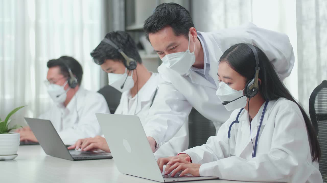 男性が女性を助けるために来るヘッドセットとマスクをかぶった3人のアジア人医師が同僚がオフィスでの通話中に話しタイピングしている間コールセンターエージェントとして働くためにコンピューターを使用しています