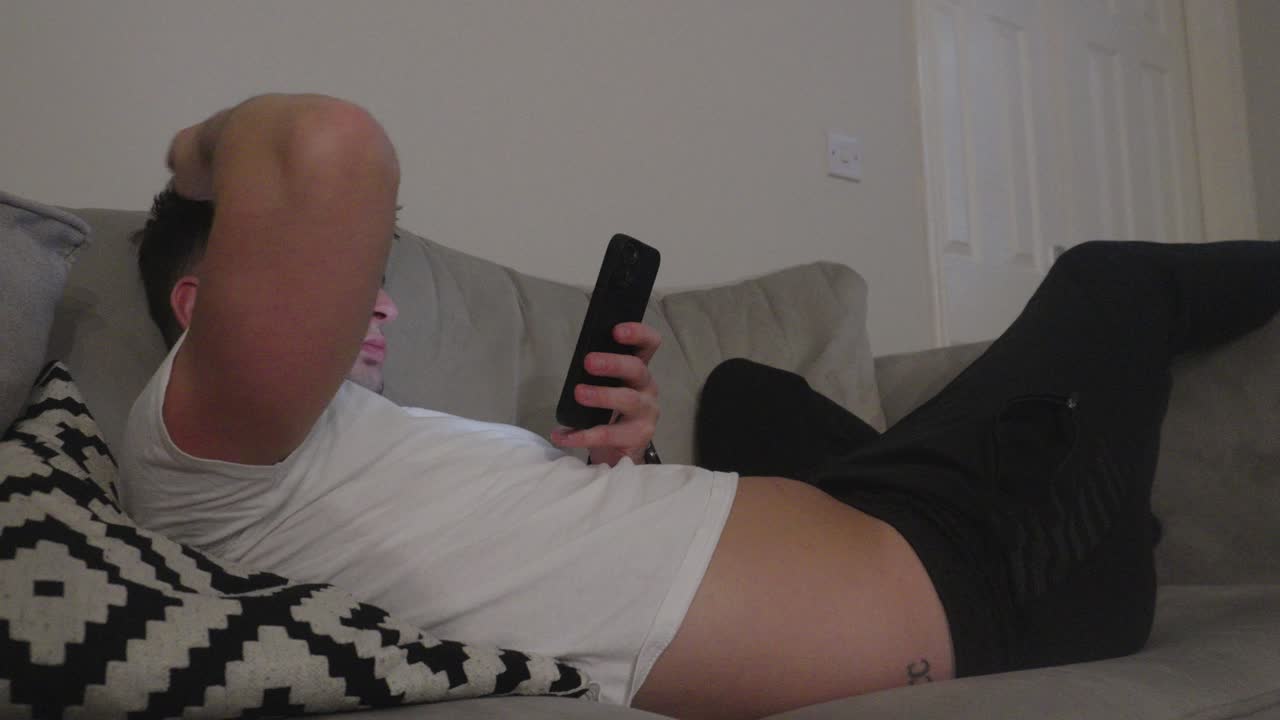 젊은 예쁜 남자가 소파에서 휴식을 취하고 스마트폰을 스크롤하고 있습니다.