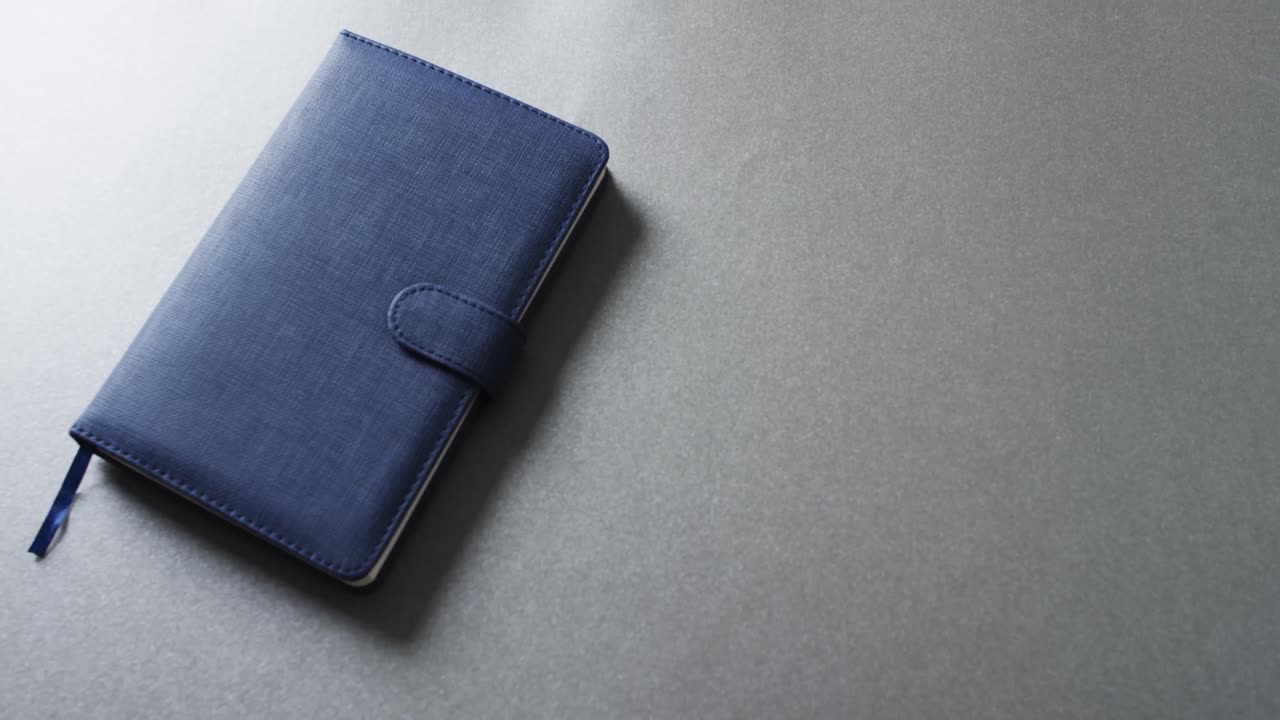 Detailní záběr zavřeného modrého zápisníku s prostorem pro text na šedém pozadí ve zpomaleném záběru.