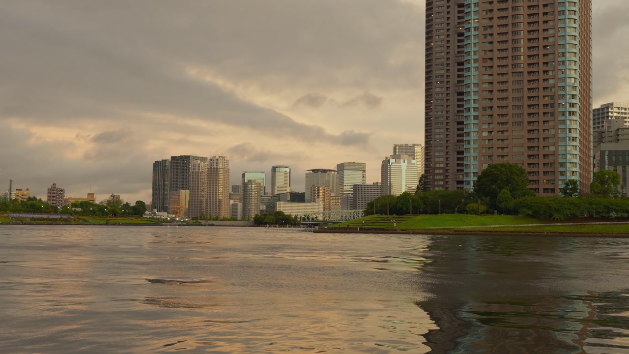 A wide shot of the Tokyo cityscape and a river, with tall buildings silhouetted against a cloudy sunset sky.