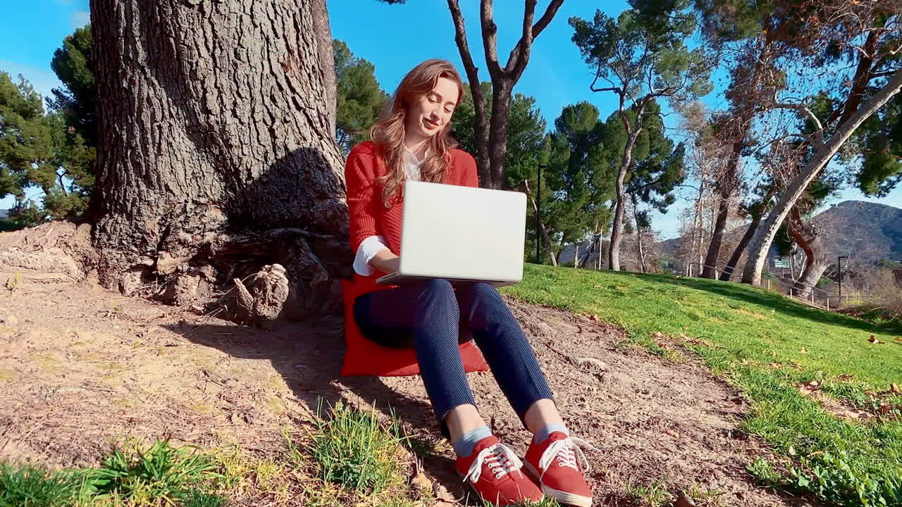 una mujer feliz, atractiva, joven y caucásica en el parque, escribiendo en una computadora portátil debajo de un árbol