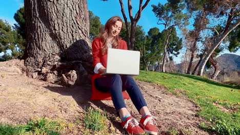 A-happy,-attractive,-young,-caucasian-woman-at-the-park,-typing-on-a-laptop-under-a-tree