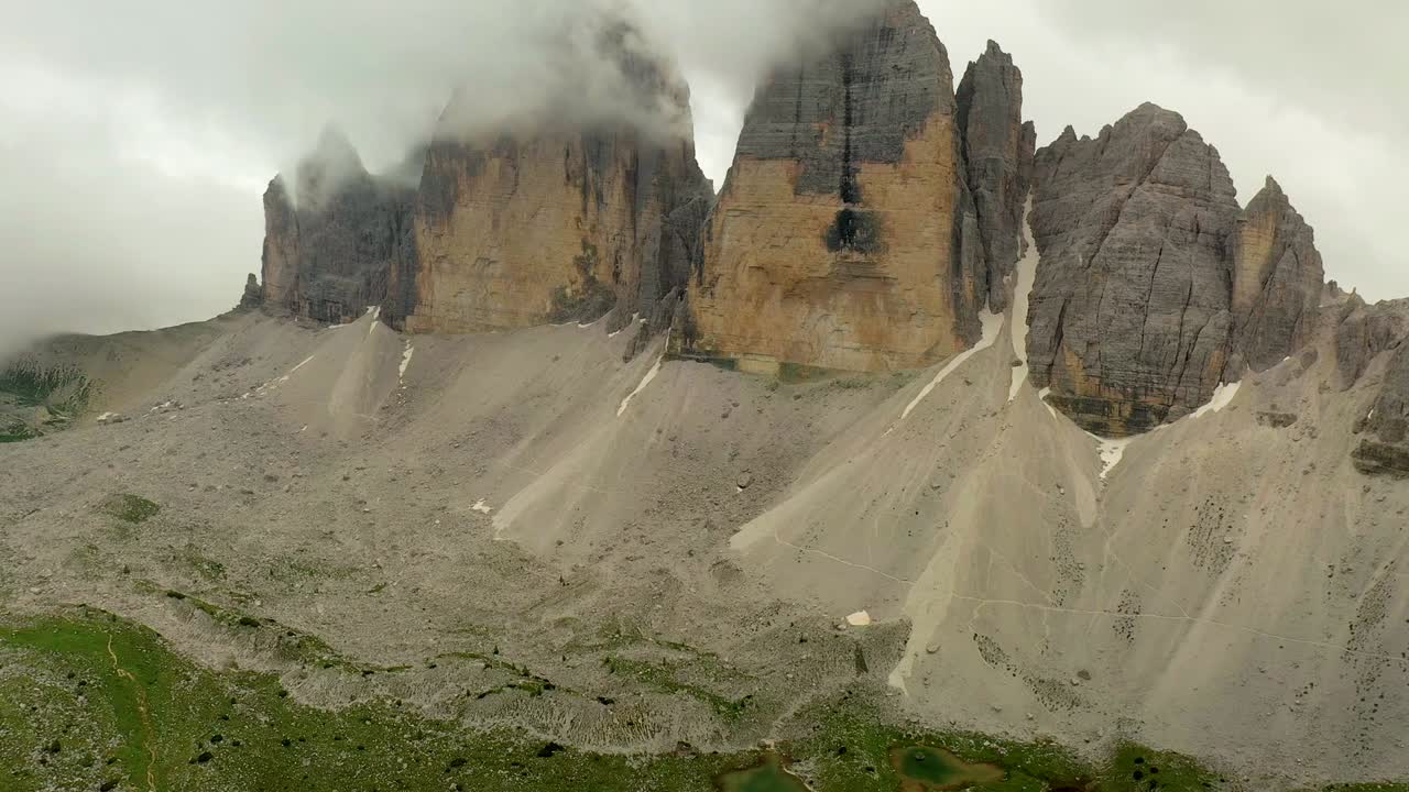 Dramatic drone view of Tre Cime di Lavaredo peaks in the Dolomites, with rocky slopes, rugged cliffs, and misty clouds drifting around the towering mountain landscape