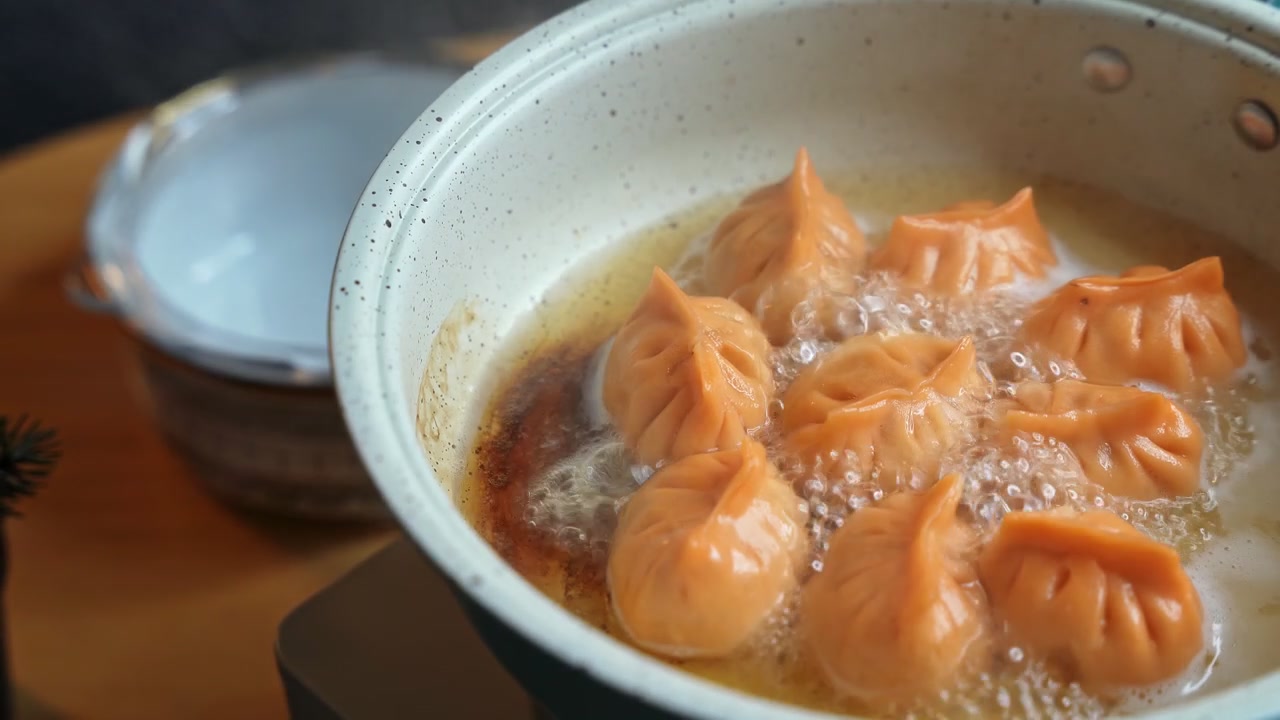 Bidikan jarak dekat pangsit oranye yang digoreng dalam minyak bening mendidih di wajan berbintik terang. Dim sum Asia yang lezat ini dimasak di meja kayu dalam suasana dapur rumah modern di dalam ruangan.