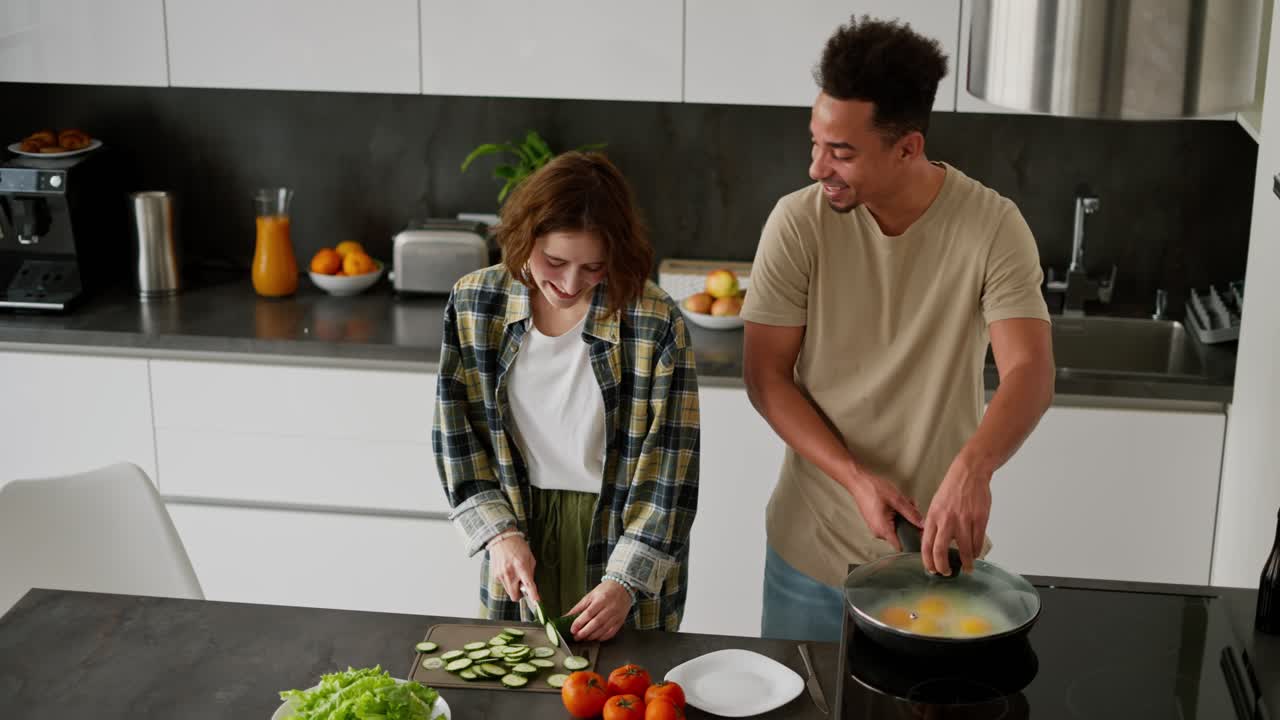 feliz joven moreno de piel negra en una camiseta de crema cocina huevos revueltos y freír su desayuno mientras su joven novia madura prepara ensalada por la mañana en un apartamento moderno en una mesa de cocina negra