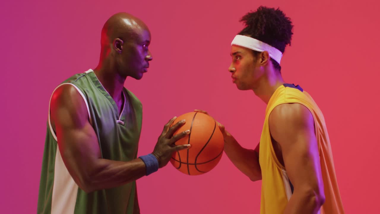 Video of two diverse male basketball players facing each other with ball on pink background