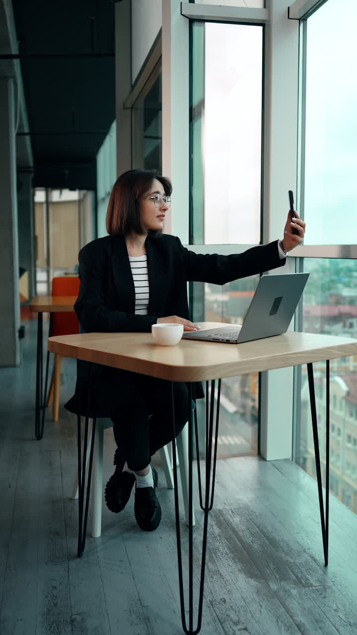 Woman in black jacket sits at desk holding her phone high in the hand. Girl taking selfie or video. Vertical video.
