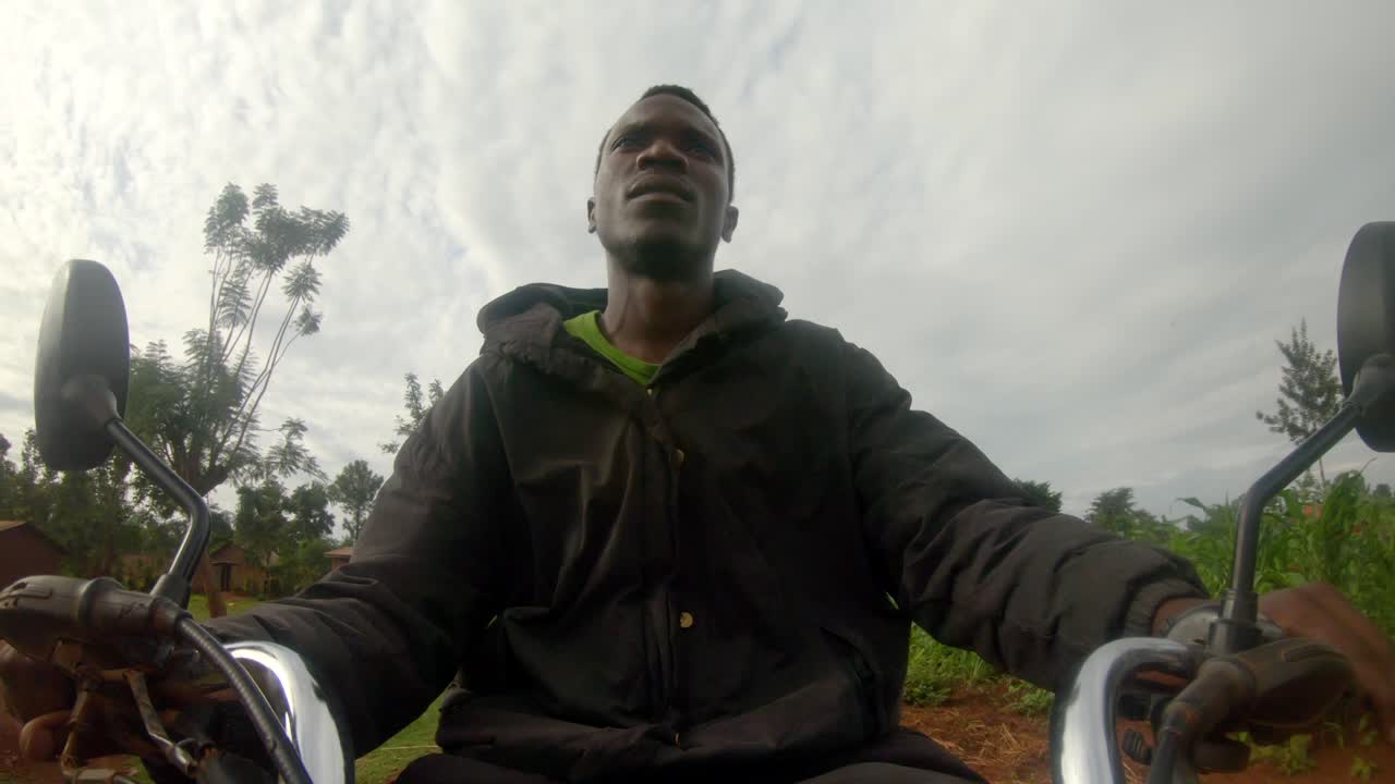 Et actionbillede af en afrikansk mands ansigt, mens han styrer en motorcykels styr og kører gennem en afrikansk landsby.