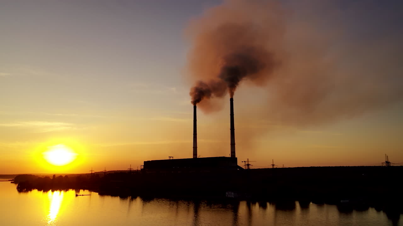 Industrial factory near the river at sunset. Smoke comes out of the pipes of a huge plant in the evening. Environmental pollution.