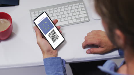 Hombre-En-El-Escritorio-Sosteniendo-Un-Teléfono-Inteligente-Con-Certificado-De-Vacunación-Covid,-Bandera-De-La-Ue-Y-Código-Qr-En-La-Pantalla