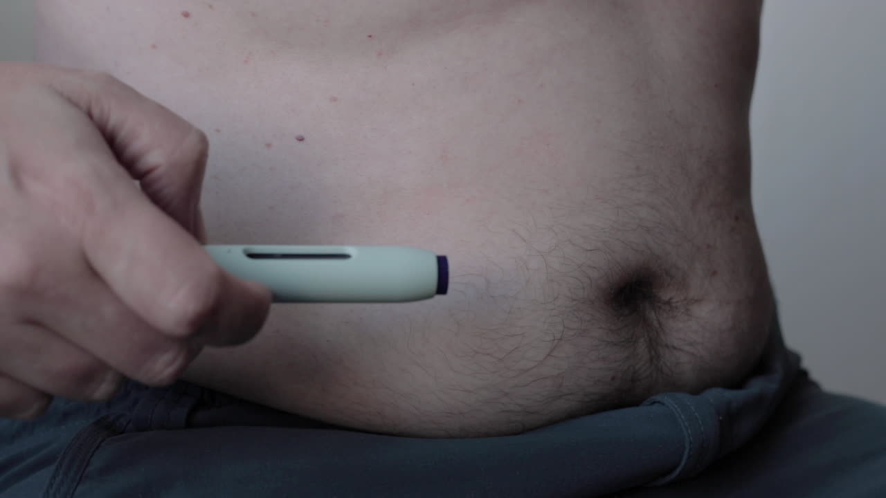 Primo piano dell'auto-somministrazione di un farmaco tramite una penna con ago medico che inietta la sostanza in un'area adiposa dell'addome, lasciando una goccia di sangue come segno
