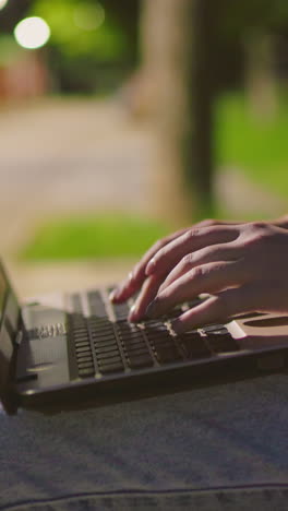 primer plano de una mujer usando una computadora portátil al aire libre por la noche, los dedos escribiendo en el teclado con reflejo blanco en la pantalla, con un fondo borroso suave iluminado