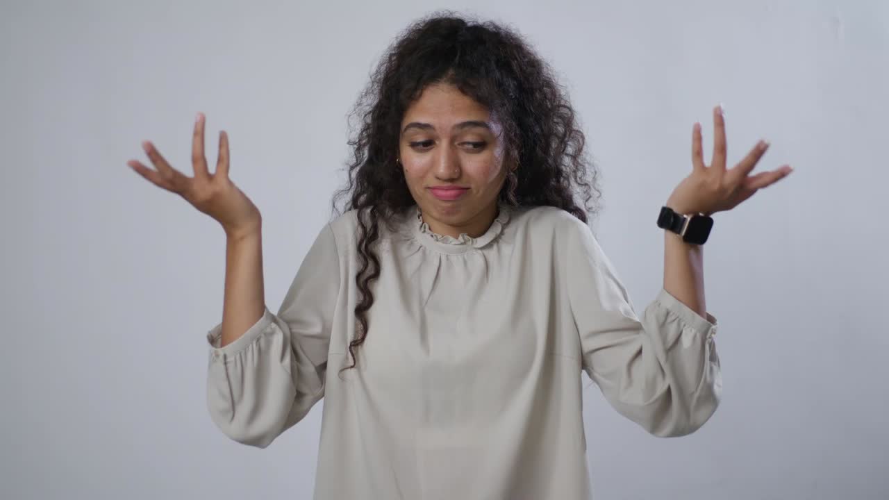 A young South Asian woman with curly brown hair points the viewers and makes an "OK" hand gesture