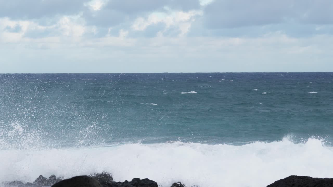 A dramatic coastal scene of a massive ocean wave colliding with volcanic rocks, creating a towering explosion of white spray and foam. The clip captures the raw power of the sea