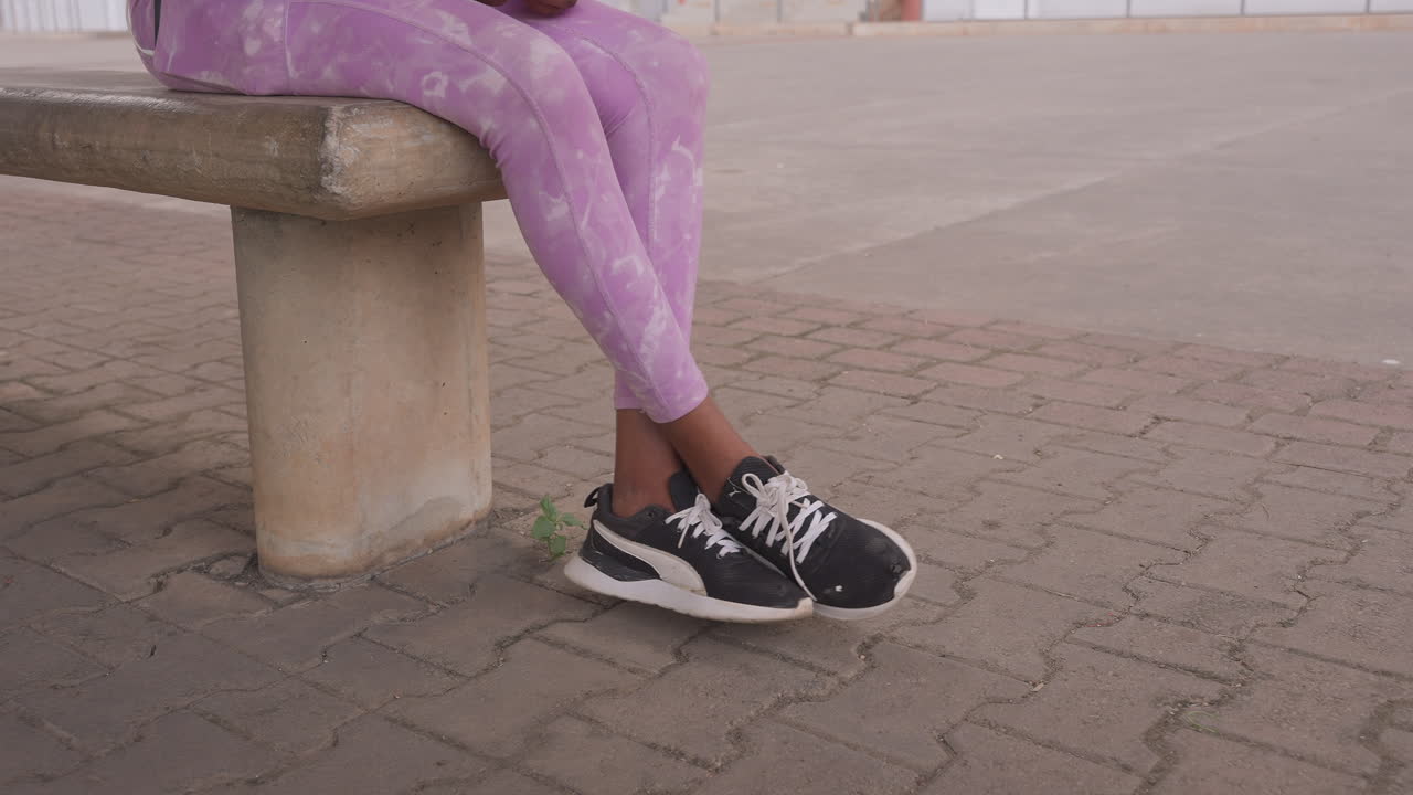 Gambe di donna nera sedute su una panchina, leggings a fantasia viola e scarpe da ginnastica bianche e nere su pavimentazione ruvida, primo piano di piedi e parte inferiore delle gambe, ambientazione di una piazza urbana in cemento, posa di attesa rilassata