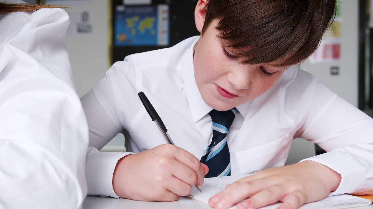 Studente delle scuole superiori in uniforme che scrive su un quaderno seduto alla scrivania in classe
