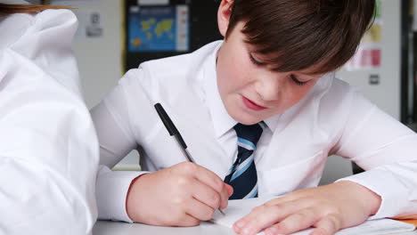 Male-High-School-Student-Wearing-Uniform-Writing-In-Exercise-Book-Sitting-At-Desk-In-Classroom