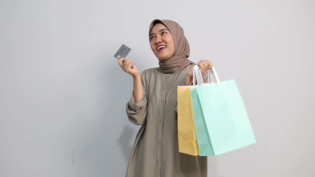 Potret wanita muslim Asia muda menunjukkan dan mempresentasikan kartu kredit atau kartu debit serta memegang tas belanja yang diisolasi dengan latar belakang putih, belanja online, konsep keuangan