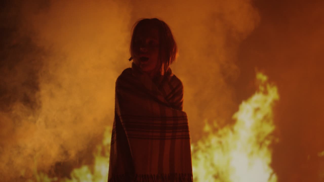 Person wrapped in blanket stands before intense fire and smoke