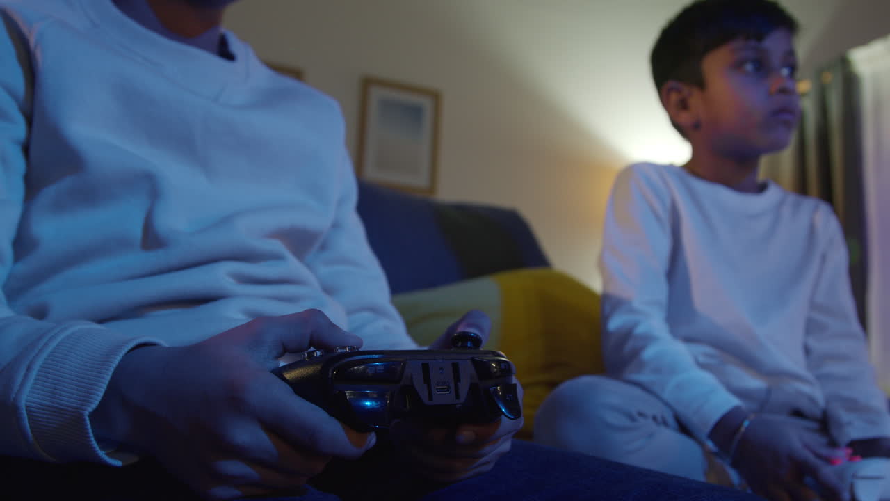 cerca de las manos de dos niños jóvenes en casa jugando con consola de juegos de computadora en la televisión sosteniendo controladores a última hora de la noche 5