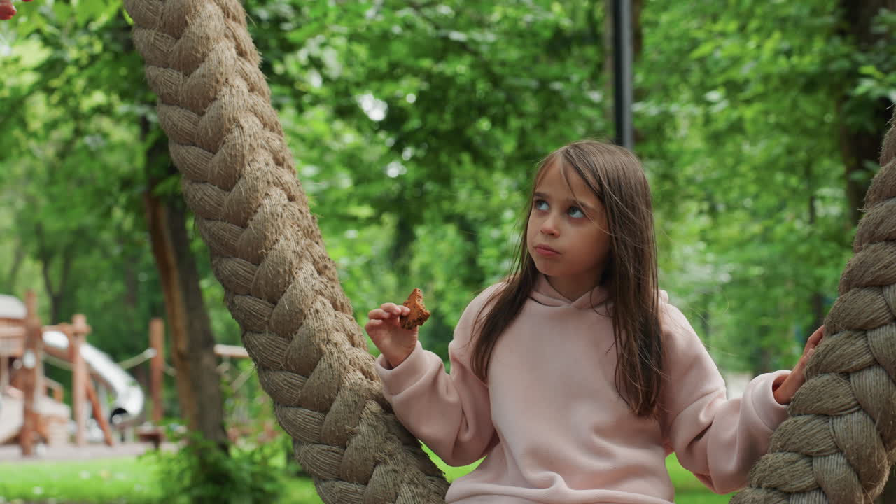 Hija pequeña en un columpio comiendo una galleta en un parque infantil bajo árboles verdes, cabello trenzado con suéter rosa, sosteniendo un dulce cerca de una cuerda gruesa, una mirada sutil hacia la mamá fuera de cuadro, un momento familiar espontáneo y contemplativo.
