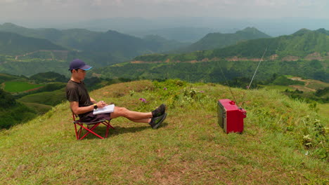 young-man-watches-television-in-a-relaxing-outdoor-atmosphere,-taking-notes-on-an-important-broadcast