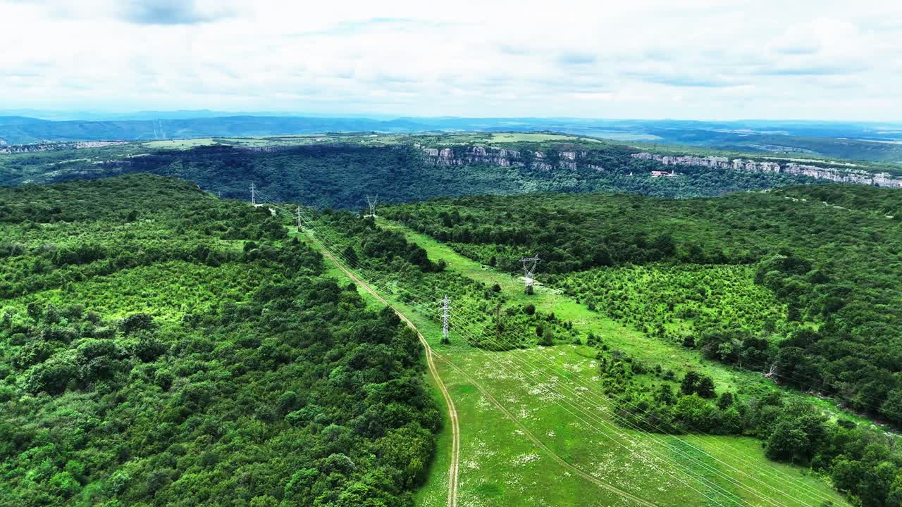 Dron se vznáší nad bujnou balkánskou zelení k impozantnímu útesu pod modrou oblohou a vzdouvajícími se mraky, s nenápadnými stopami venkovských cest vespod.