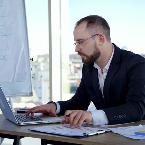 Serious male worker using laptop computer. Focused businessman working on laptop in office with large windows. Businessman in glasses typing on a computer