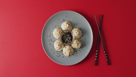 Composition-of-plate-with-dim-sum-dumplings-and-chopsticks-with-soy-sauce-on-red-background