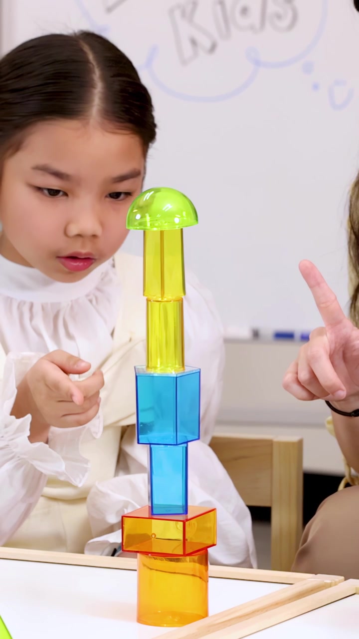 Young girl carefully builds tall tower with colorful translucent blocks in bright classroom setting