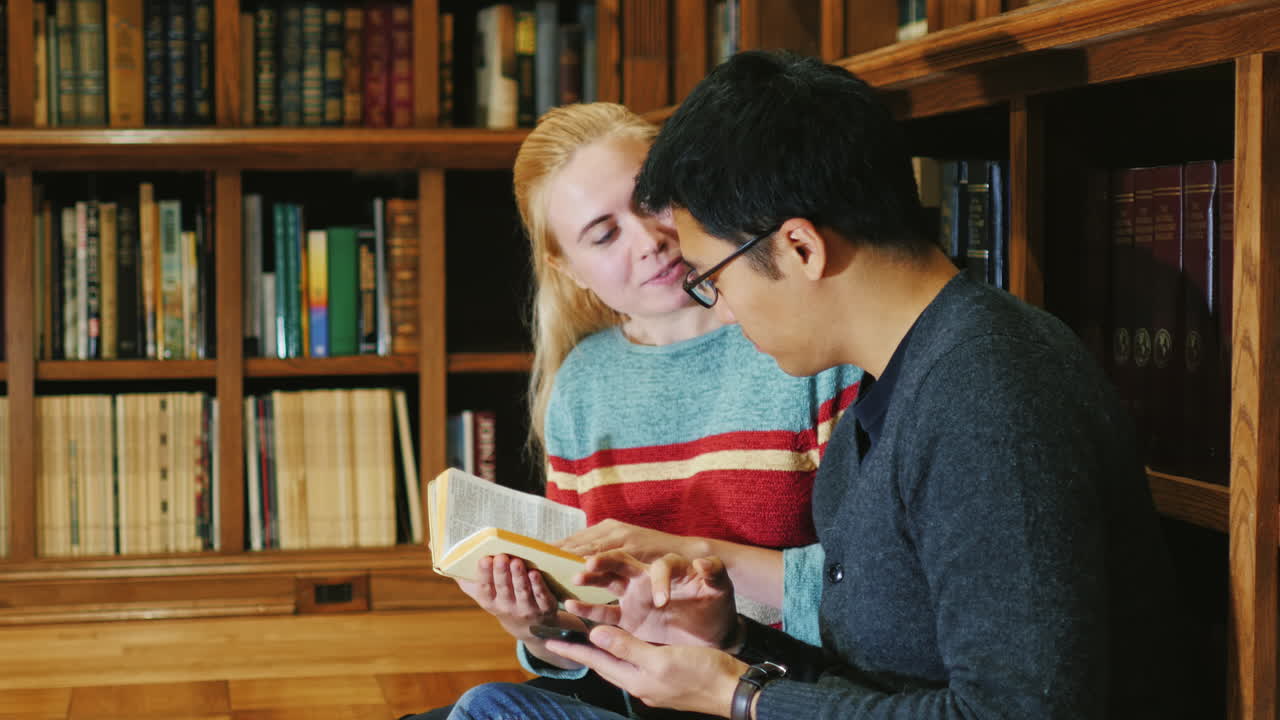 el hombre coreano y la mujer caucásica miran un libro en la biblioteca se comunican usando una tableta