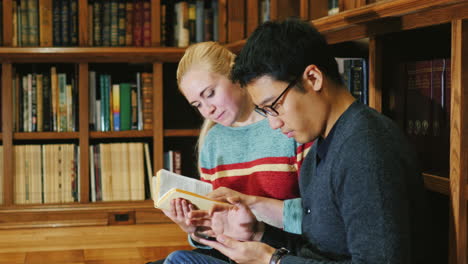 Korean-Man-And-Caucasian-Woman-Look-At-A-Book-In-The-Library-Communicate-Using-A-Tablet