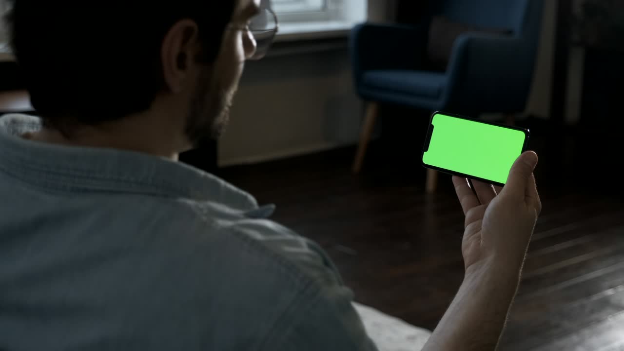 retrato en primer plano de un joven con gafas mirando un teléfono inteligente, aplicaciones en línea, redes sociales, videos y fotos, tendido en un sofá en una habitación en casa