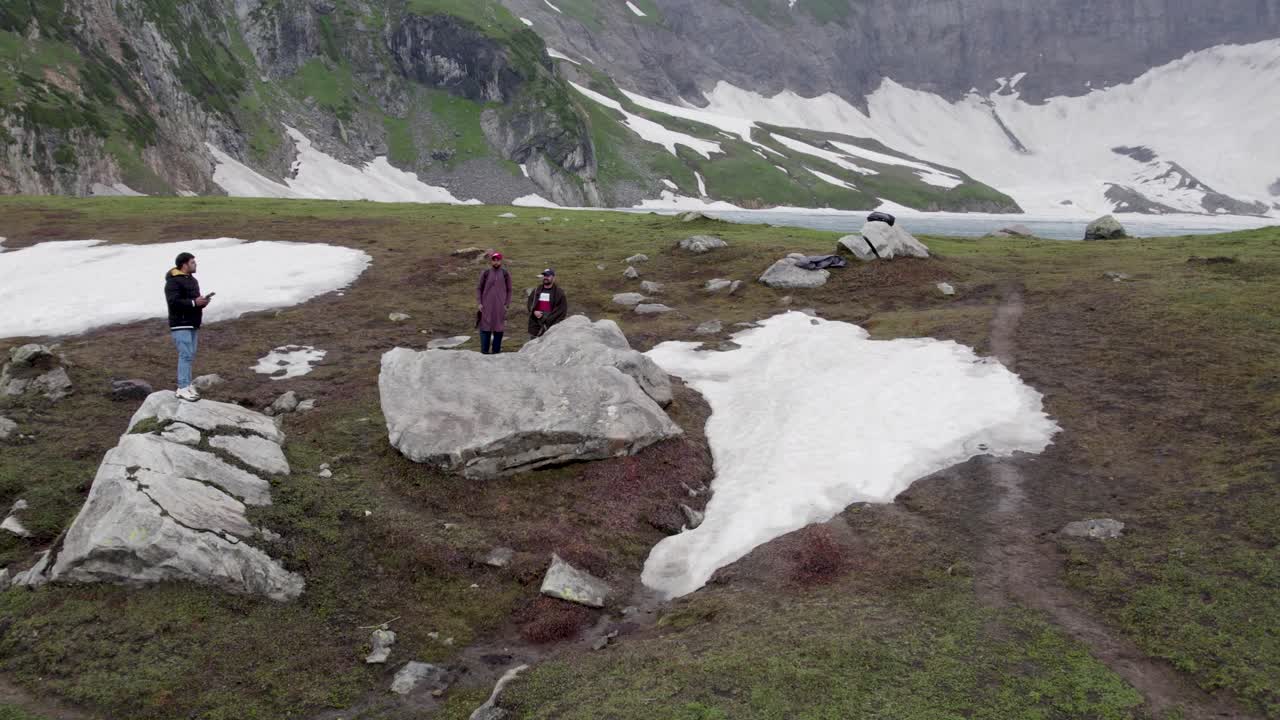 Hikers resting on rocks near snow patches in the green alpine terrain by Ratti Gali Lake