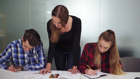 Three-students-working-on-their-homework-sitting-together-at-the-table-while-one-girl-is-dictating-and-two-other-students-are-writing.-Slowmotion-shot