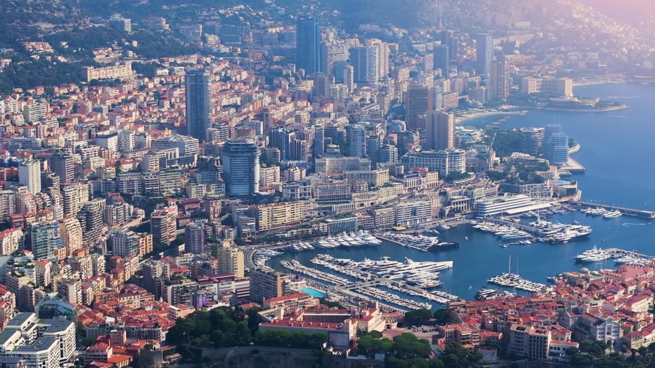La costa di Monaco presenta edifici, barche e acque limpide. Le vedute includono yacht nel porto e residenze raggruppate sulle colline. La luce del primo mattino esalta il paesaggio.