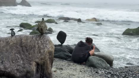 unrecognizable female in a black hoodie tediously balancing rocks on a beach in dana point california while waves crash beside her on a foggy day