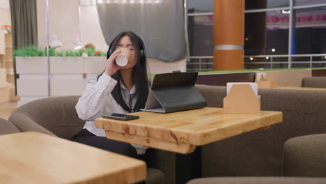 secretary in white shirt with long hair and headphones seated in cozy restaurant lounge working on tablet with focused expression while takeaway coffee and phone rest on table