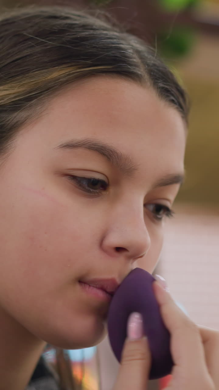 Applying makeup attentively, Careful sponge dab for natural look, Focused application of blush and lip tint, Teenager meticulously dabbing sponge on lips and cheeks in soft indoor light