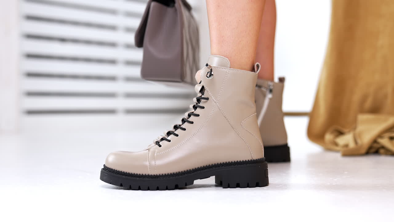 Beige leather boots with black laces and tractor soles on model's feet. Close up. Grey bag hanging at backdrop. Studio shot.