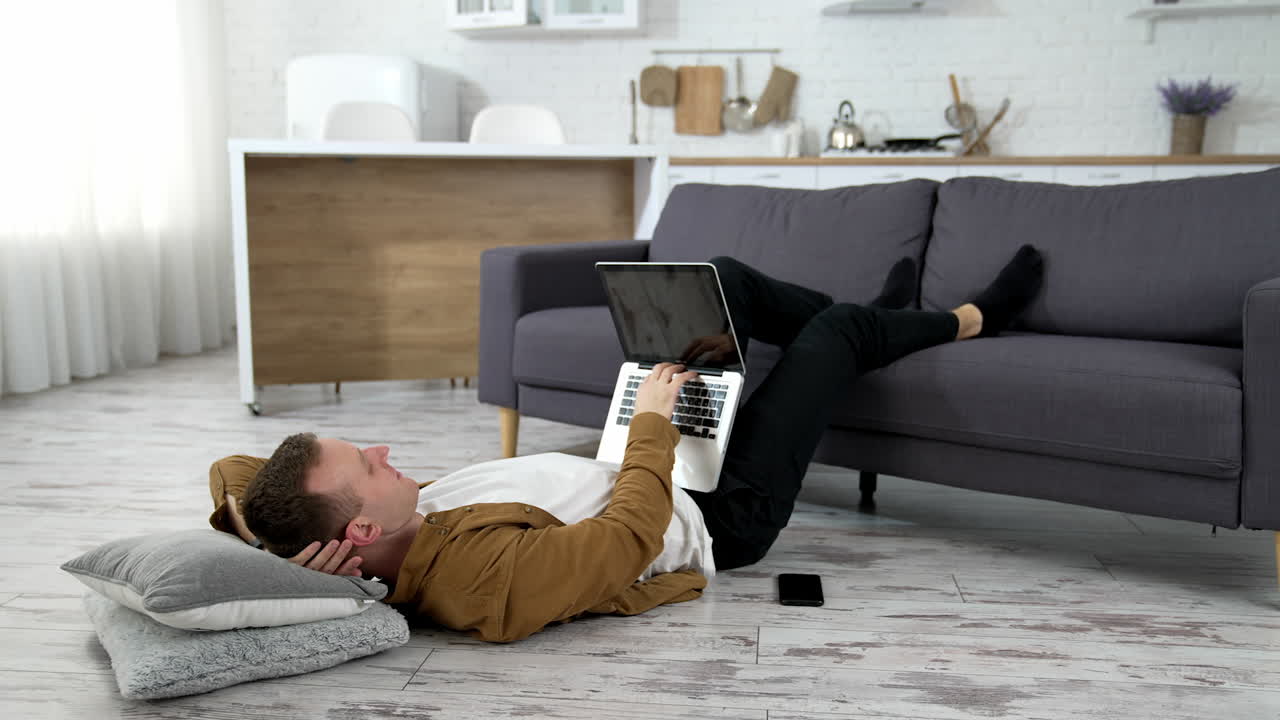 Freelancer working at home. Tired student studying on a laptop while laying on a floor with his legs on sofa. Remote job.