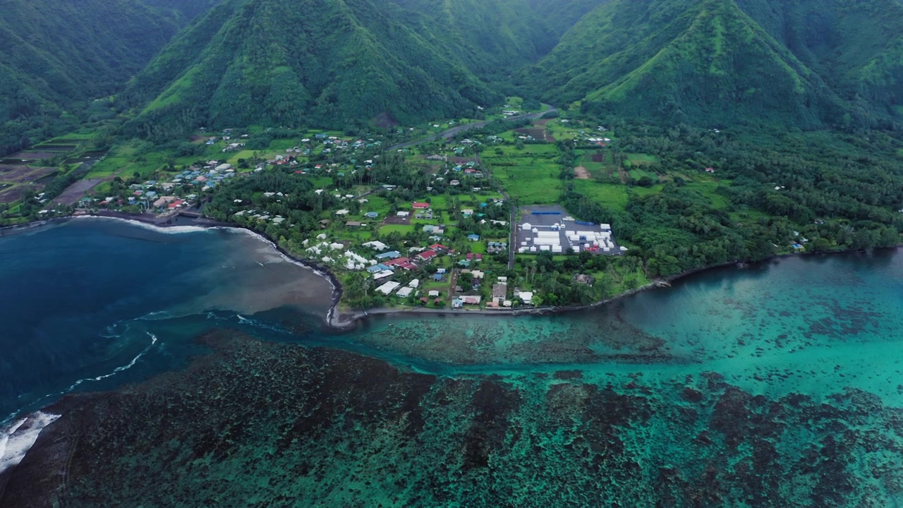 Vista aerea con drone del villaggio di Teahupoo, Tahiti, adagiato sotto ripide montagne della foresta pluviale mentre un'onda si infrange lungo la barriera corallina di fronte alla costa