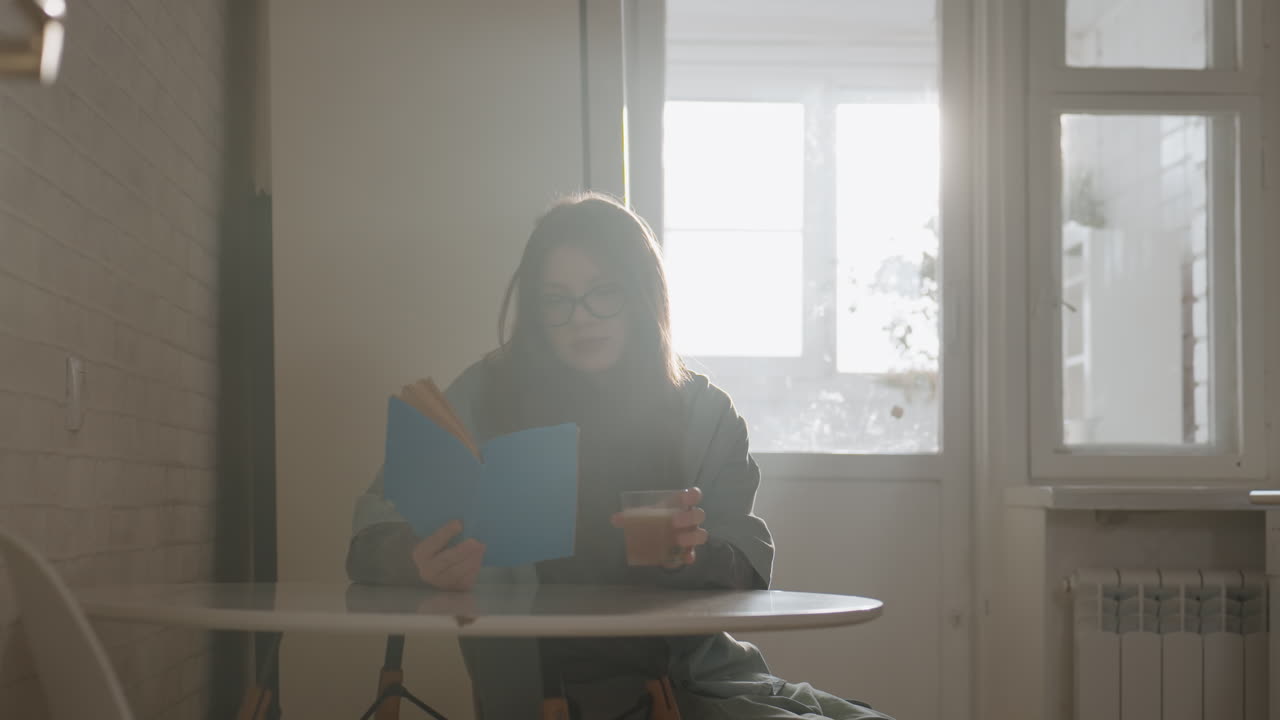 Giovane donna che legge vicino alla finestra, sorseggiando caffè, sfogliando le pagine di un quaderno blu, luce solare calda che filtra dalla finestra, bagliore dell'obiettivo morbido, interno accogliente di un appartamento con tavolo e sedia, occhiali appoggiati