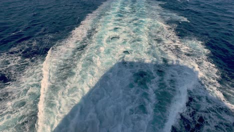 back view of wide water wake left from cruise ship on sea water surface with horizon in background and shadow of people leaning over parapet of deck