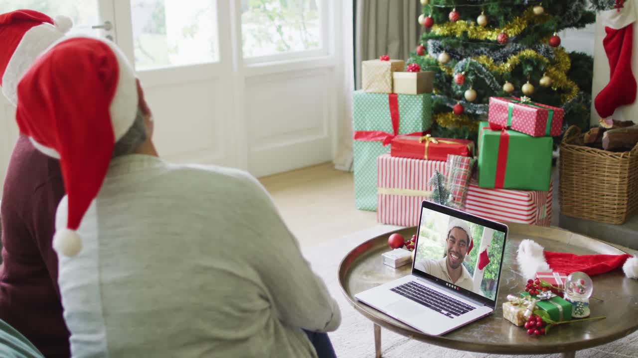 amigas mayores diversas usando una computadora portátil para una videollamada de navidad con un hombre sonriente en la pantalla