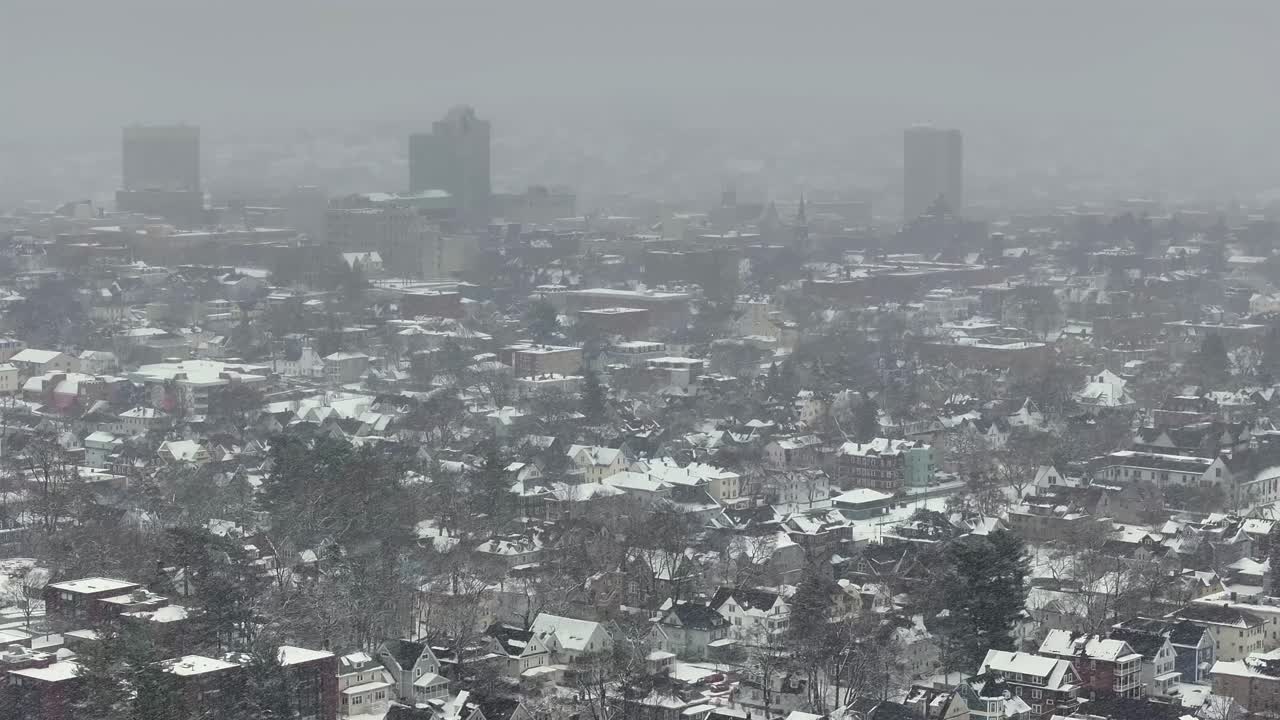 Downtown Worcester towers loom behind snow-clad neighborhoods. Houses crowd the hillside. Sweeping across city rooftops, aerial.