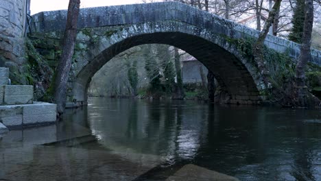 Vista-En-ángulo-Bajo-De-Pasos-De-Aguas-Poco-Profundas-Hacia-El-Río-Molgas-Y-Un-Puente-Romano-Con-Arco