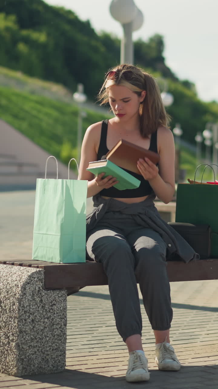 dama sentada en un banco al aire libre con bolsas de compras a ambos lados, recuperando un libro de una bolsa verde menta y colocando un libro marrón de nuevo en la misma bolsa, el fondo incluye postes de lámpara